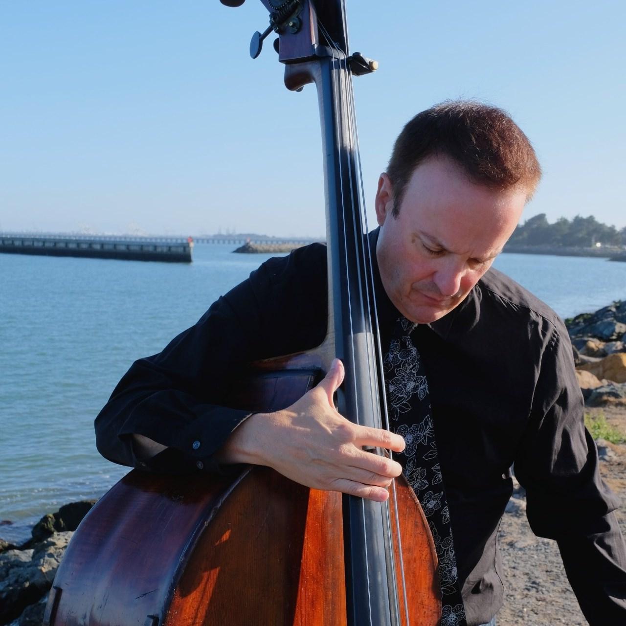 Michel Taddei, in a white dress shirt, holding a double bass. He looks down and sits with his back against the Bay