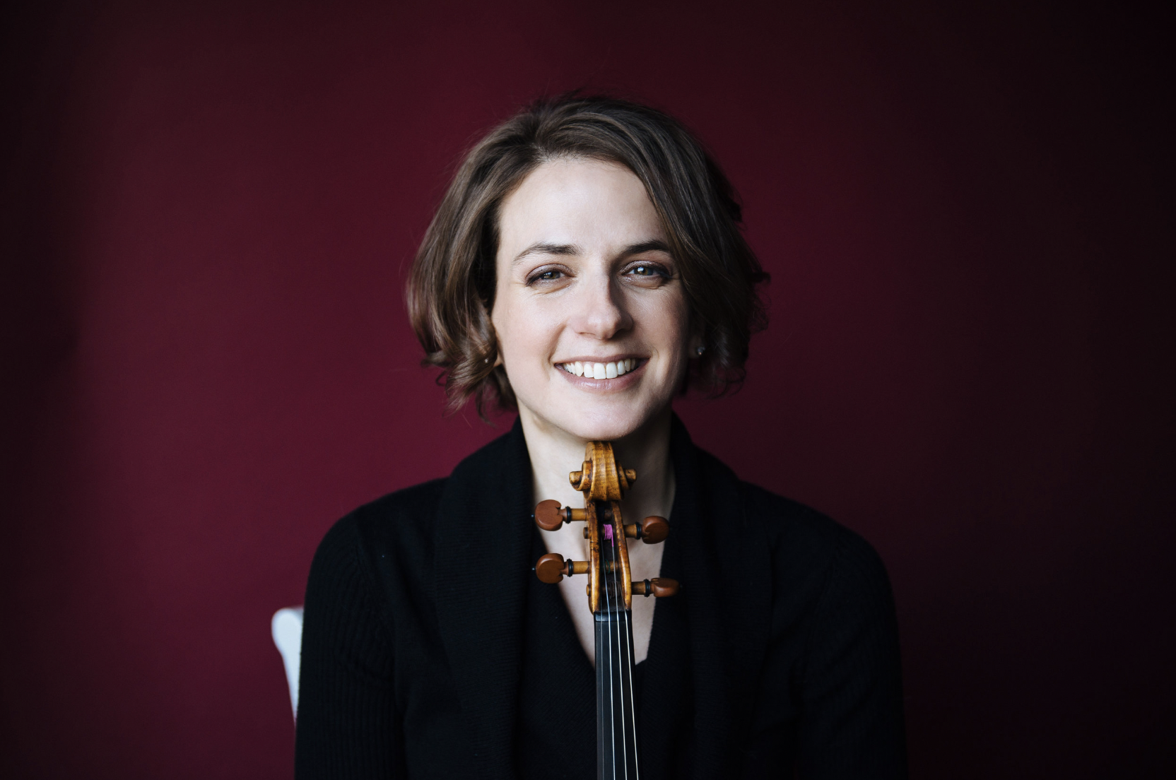 Liana Bérubé against a red background, smiling, and resting her chin on a violin