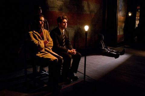 两个人坐着，被一个灯泡照亮. 他们的环境(舞台)沉闷而黑暗. 在角落里，一个男人坐在地板上，躲在阴影里.