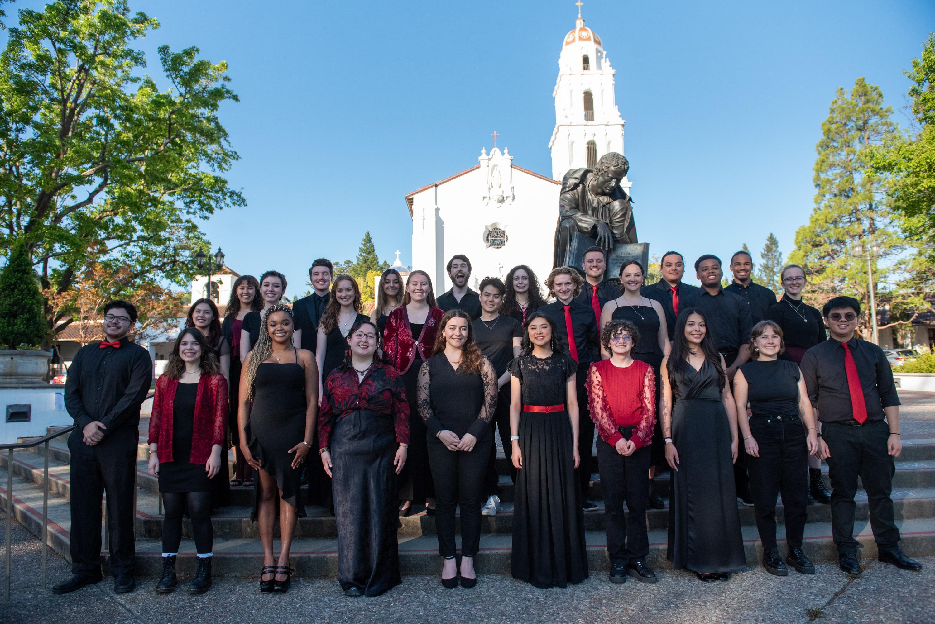 Saint Mary's College choir dressed in black and red stands in front of the SMC Chapel and statue of John Baptist de la Salle with a blue sky above them.