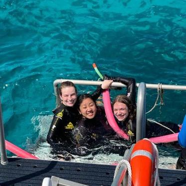 三名学生穿着潜水装备，在明亮的绿松石水里挂在船上拍照.