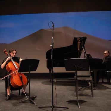 Two musicians, one piano player and one cellist, 在勒费弗剧院的舞台上表演，背景是黄昏时的沙漠景观. 屏幕中间有一个很大的沙丘. 