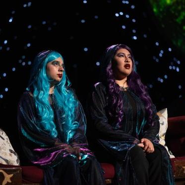 把它送上月球——两个紫色和蓝色头发的女人从舞台后面望出去, 有一束光照在他们身上. 在他们身后，是宇宙的背景. 