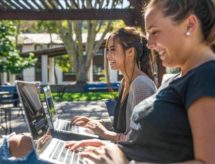 Two students laughing while on their laptops outside